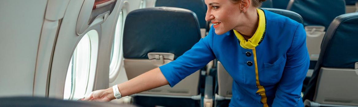 Joyful woman flight attendant in air hostess uniform opening aircraft window and smiling while standing near passenger seats