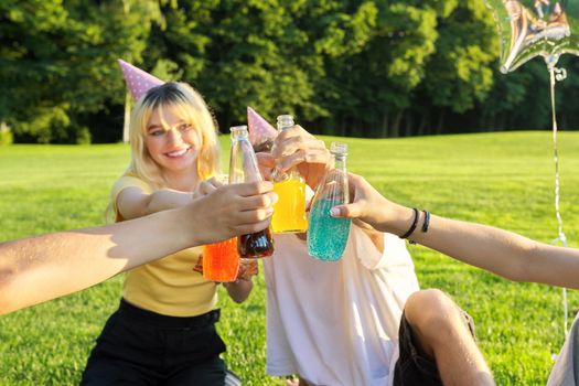 Teenage birthday party picnic on grass in park. Group of teenagers having fun, congratulating girl on her 17th birthday, drinking colored fruit and soft drinks. Age, adolescence, fun, holiday concept