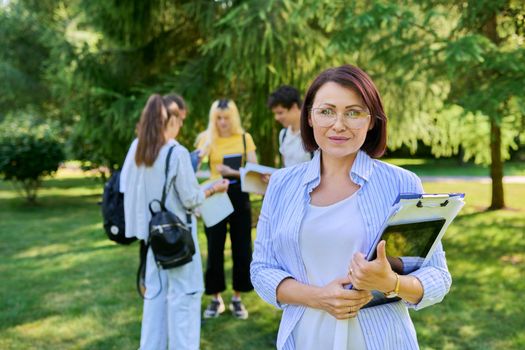 Middle aged female teacher mentor in glasses with digital tablet looking at camera, group of teenage students in park school college background. Education, teaching, profession, teachers day concept