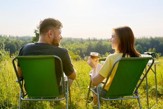 Happy middle aged couple relaxing together outdoor sitting on folding chairs at camping table. Man and woman talking, drinking fresh coffee, enjoying summer nature of wild meadow, back view, weekend