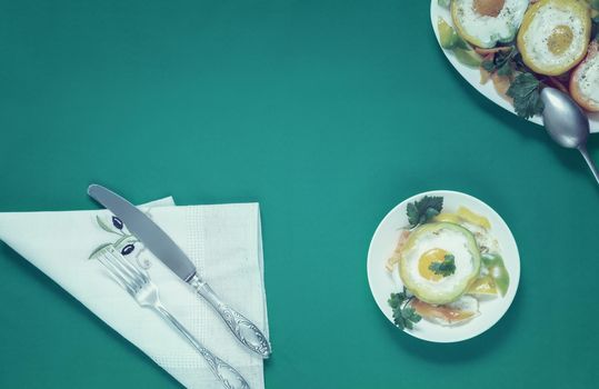 On the table on plates are baked in the oven bell peppers with an egg inside each pepper. There are Cutlery on a napkin nearby. Top view with space to copy. Flat lay