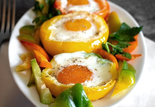 On the table on plates are baked in the oven bell peppers with an egg inside each pepper. There are Cutlery on a napkin nearby. Front view, close-up.