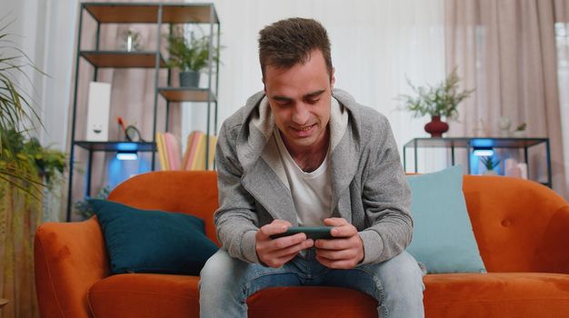 Worried funny young adult man enthusiastically playing shooter online video games on smartphone. Excited player boy using mobile phone gadget app celebrate win victory at home sitting on couch alone