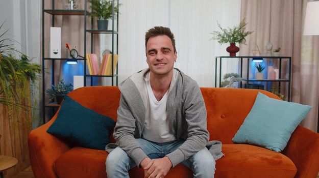 Portrait of happy one handsome teenager unshaved guy in casual shirt smiling, looking at camera. Young cute fashion model man indoors isolated at home in living room on couch. Male nature beauty