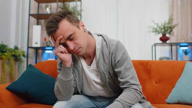 Portrait of sad young Caucasian man sitting at home looks pensive thinks over life concerns or unrequited love, suffers from unfair situation. Problem, break up, depressed feeling bad annoyed, burnout