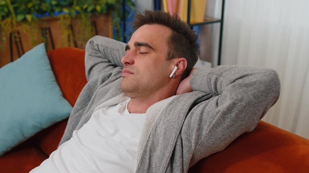 Happy young man wearing wireless earphones listening favorite energetic disco music song, relaxing, sleeping, resting, having a break, lying on couch at home. Weekends entertaining activities indoors