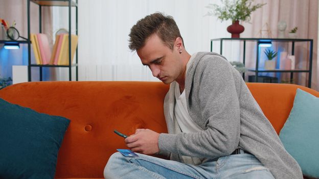 Smiling Caucasian young adult man sitting on couch using credit bank card and smartphone while transferring money, purchases online shopping in living room home. Male guy tapping, scrolling, browsing