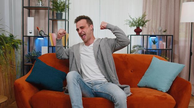Handsome happy guy in casual t-shirt shouting, raising fists in gesture I did it, celebrating success, winning and goal achievemen, receive good news. Young man indoor at home in living room on couch