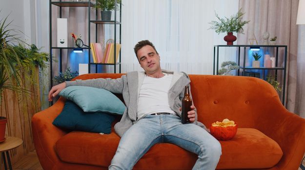 Excited young man sitting on sofa eating chips, drinking beer and watching interesting tv serial, football championship game, social media content online at home. Guy enjoying domestic entertainment