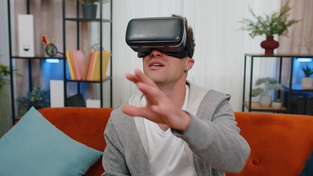 Excited man wearing VR headset resting at home sitting on sofa in living room alone using futuristic technology making gestures with hands in air, play 3D video game movie. Virtual reality glasses