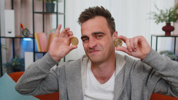 Man freelancer holding two gold coins at home, successful developer programmer. Guy stock trader earning bitcoins after online monitoring trading operations. Increasing wealth, financial prosperity