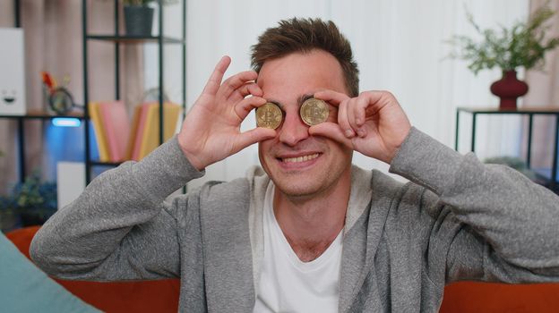 Man freelancer holding two gold coins at home, successful developer programmer. Guy stock trader earning bitcoins after online monitoring trading operations. Increasing wealth, financial prosperity