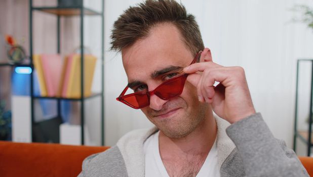 Portrait of seductive cheerful stylish unshaved successful man wearing red sunglasses, charming smile, blinking, winking eye, flirting. Young lovely adult guy sitting at home in living room on couch