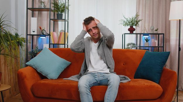 Repair work at neighbours concept. Irritated man sit on couch cover ears with hands annoyed by noisy neighbors suffers from headache wishes silence. Thin walls at home, flat without sound insulation