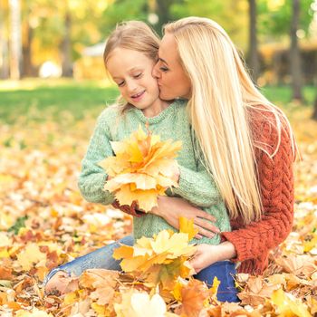 Happy young caucasian mother and little daughter holding autumn yellow leaves sitting and kissing at the park