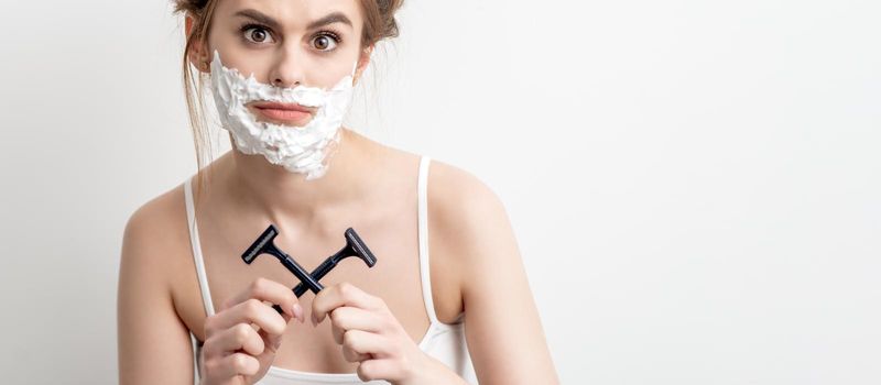 Beautiful young caucasian woman with shaving foam on her face and two razors in her hands on white background