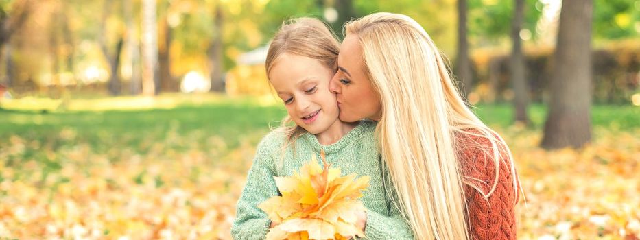 Happy young caucasian mother and little daughter holding autumn yellow leaves sitting and kissing at the park