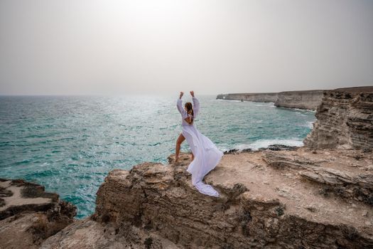 Happy freedom woman on the beach enjoying and posing in white dress over the sea. View of a girl in a fluttering white dress in the wind. Holidays, holidays at sea