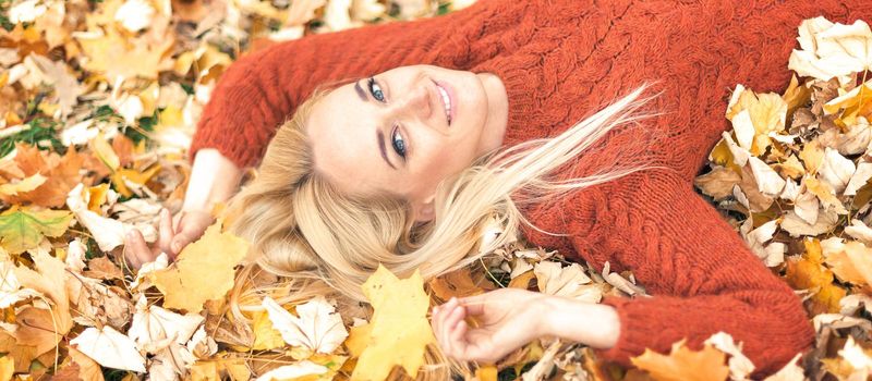 Beautiful young caucasian woman lying down on yellow leaves looking at camera in Autumn Park