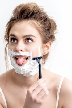 Beautiful young caucasian woman shaving her face by razor on white background. Pretty woman with shaving foam on her face