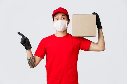 Concept of delivery and carriers during coronavirus pandemic. Asian courier in red uniform, medical mask and protective gloves working on quarantine, holding box package and point left.