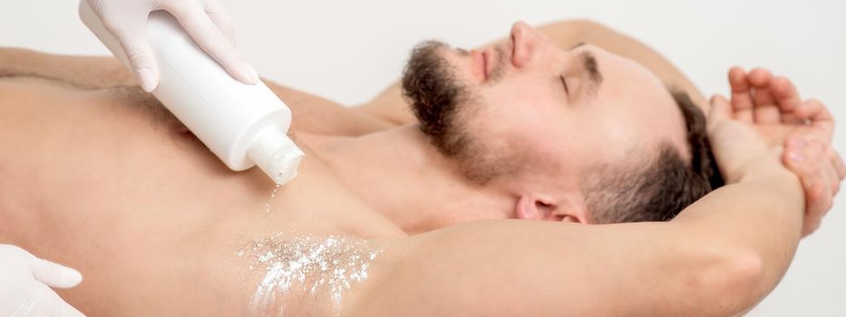 Hand of master depilation pouring talcum powder on armpit of young man before depilation procedure.