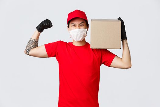 Concept of delivery during coronavirus pandemic. Courier show-off his strength, flex biceps and carry heavy package on shoulder, wear medical mask and gloves for safety on covid-19 quarantine.