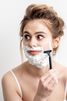 Beautiful young caucasian woman shaving her face by razor on white background. Pretty woman with shaving foam on her face