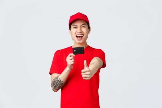 Excited smiling asian delivery guy in red cap, t-shirt uniform, showing thumb-up and credit card, recommend client great offer, promo for courier service, online shopping and internet stores concept.