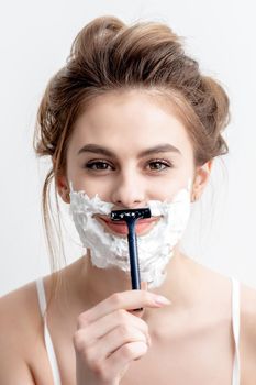 Beautiful young caucasian woman shaving her face by razor on white background. Pretty woman with shaving foam on her face