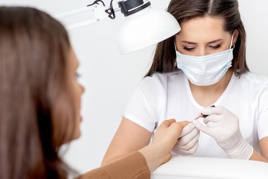 Manicurist in protective mask painting female nails with nail polish in manicure salon.