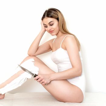 Beautiful young caucasian woman shaves her legs with a knife on a white background