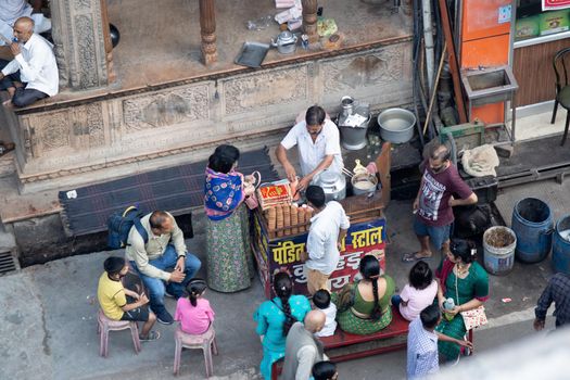 Hadiwar, Uttarakhand, India - circa 2021: aerial drone shot of road side tea seller in white shirt kurta with small stall preparing tea on a stove and old utensils by putting in milk, leaves, sugar and more as the crowds move around him in the busy city