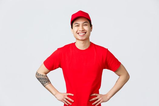 Express delivery, shipping and logistics concept. Smiling charismatic asian male delivery man in red cap, t-shirt, hold hands on hips, looking pleased, ready help client, give out customer packages.