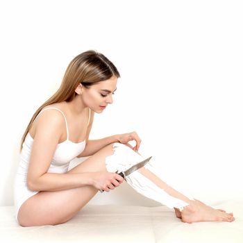 Beautiful young caucasian woman shaves her legs with a knife on a white background