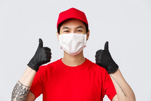 Have nice day clients. Cheerful asian delivery man using medical masks and gloves to prevent covid-19 spread and provide safe shopping or order delivery. Courier thumb-up recommend package shipping.