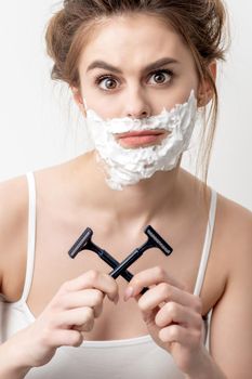 Beautiful young caucasian woman with shaving foam on her face and two razors in her hands on white background