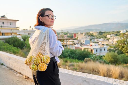 Eco tourism, woman walking relaxing in European mountain village, enjoying clean air, landscape, sunset, with a bag of fresh farm oranges. Nature, travel, ecology, lifestyle, people concept