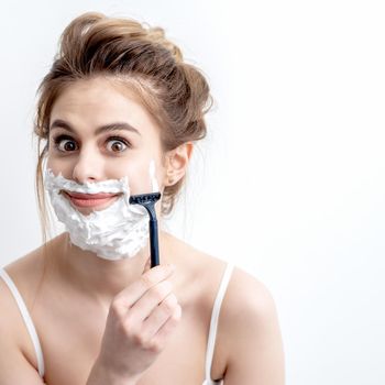 Beautiful young caucasian woman shaving her face by razor on white background. Pretty woman with shaving foam on her face