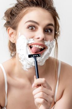Beautiful young caucasian woman shaving her face by razor on white background. Pretty woman with shaving foam on her face