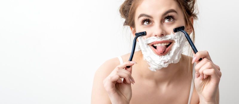 Beautiful young caucasian woman shaving her face by two razors with tongue out on white background.