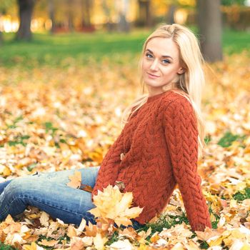 Beautiful young caucasian woman in red sweater sitting on the leaves in the autumn park