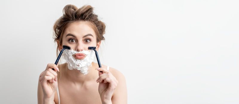 Beautiful young caucasian woman shaving her face by two razors on white background. Pretty woman with shaving foam on her face