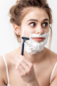 Beautiful young caucasian woman shaving her face by razor on white background. Pretty woman with shaving foam on her face