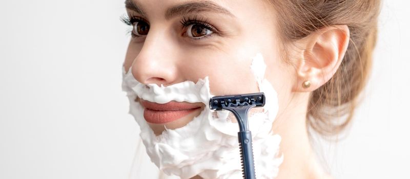 Beautiful young caucasian woman shaving her face by razor on white background. Pretty woman with shaving foam on her face