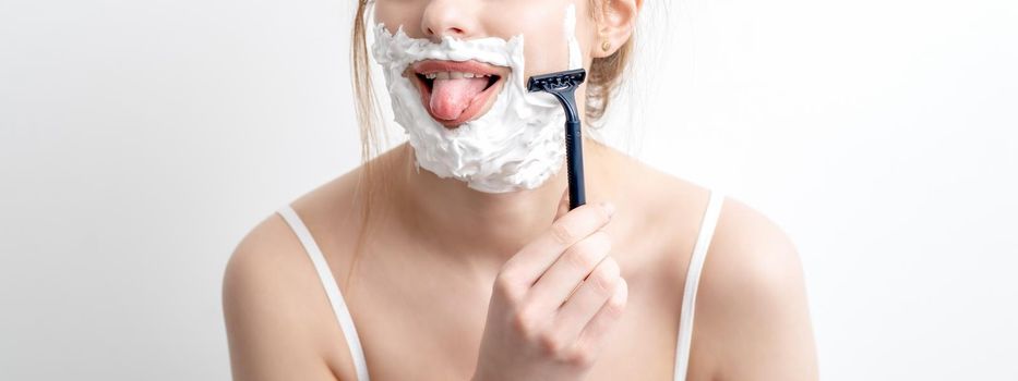 Beautiful young caucasian woman shaving her face by razor on white background. Pretty woman with shaving foam on her face