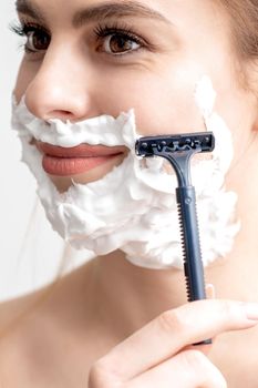 Beautiful young caucasian woman shaving her face by razor on white background. Pretty woman with shaving foam on her face