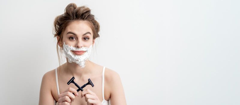 Beautiful young caucasian woman with shaving foam on her face and two razors in her hands on white background