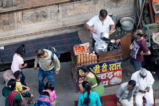 Hadiwar, Uttarakhand, India - circa 2021: aerial drone shot of road side tea seller in white shirt kurta with small stall preparing tea on a stove and old utensils by putting in milk, leaves, sugar and more as the crowds move around him in the busy city