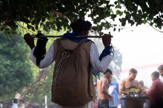 Haridwar, Uttarakhand, India - circa 2021 : bnajara wanderer with stick and backpack with turban walking along river side ghat stopping to pick leaves from a tree as the crowd around him goes about their business